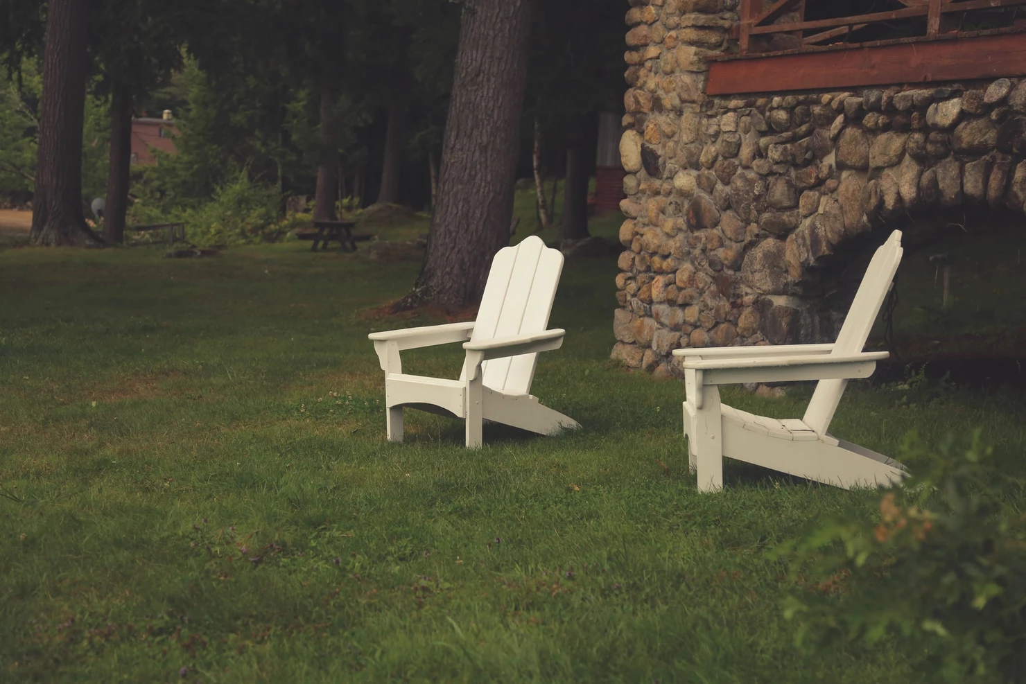 Two white Adirondack chairs on a lawn near a stone wall and picnic area